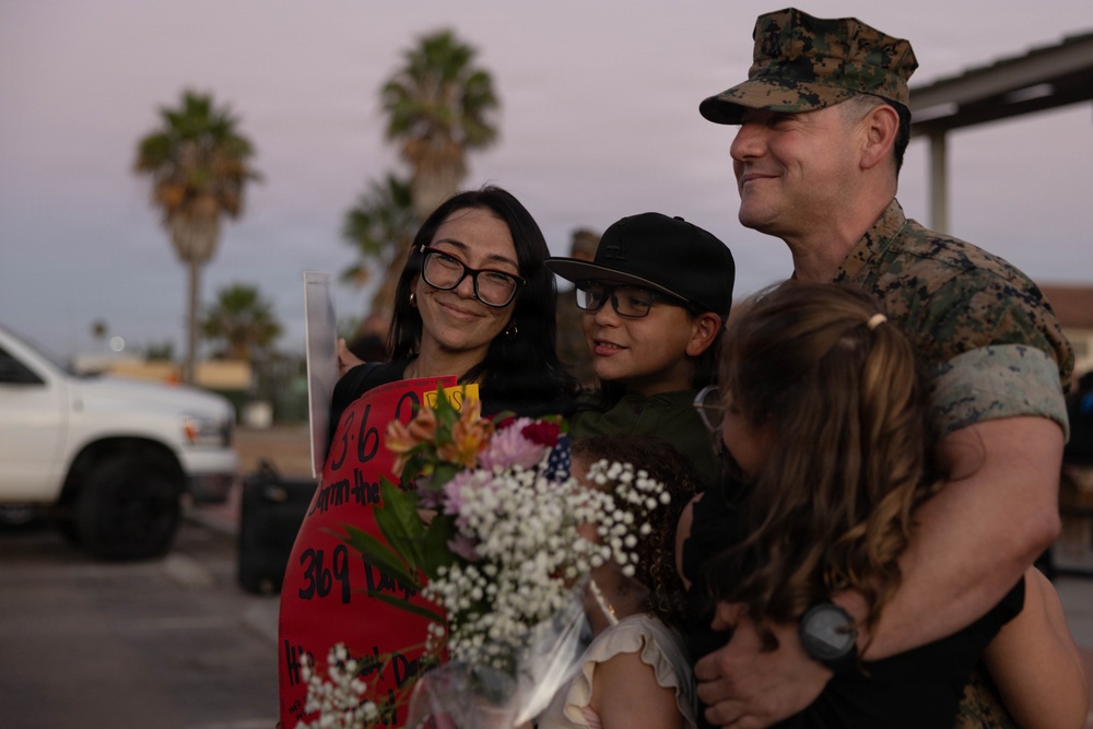 A U.S. Marine in camouflage uniform is reuniting with his family, which includes a woman holding a welcome-home sign and flowers, a boy wearing a black cap, and a young girl. The sign appears to reference '369 days,' suggesting the service member has returned from a lengthy deployment. The scene takes place outdoors at dusk, likely at a military base, with palm trees visible in the background.