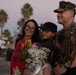 A U.S. Marine in camouflage uniform is reuniting with his family, which includes a woman holding a welcome-home sign and flowers, a boy wearing a black cap, and a young girl. The sign appears to reference '369 days,' suggesting the service member has returned from a lengthy deployment. The scene takes place outdoors at dusk, likely at a military base, with palm trees visible in the background.