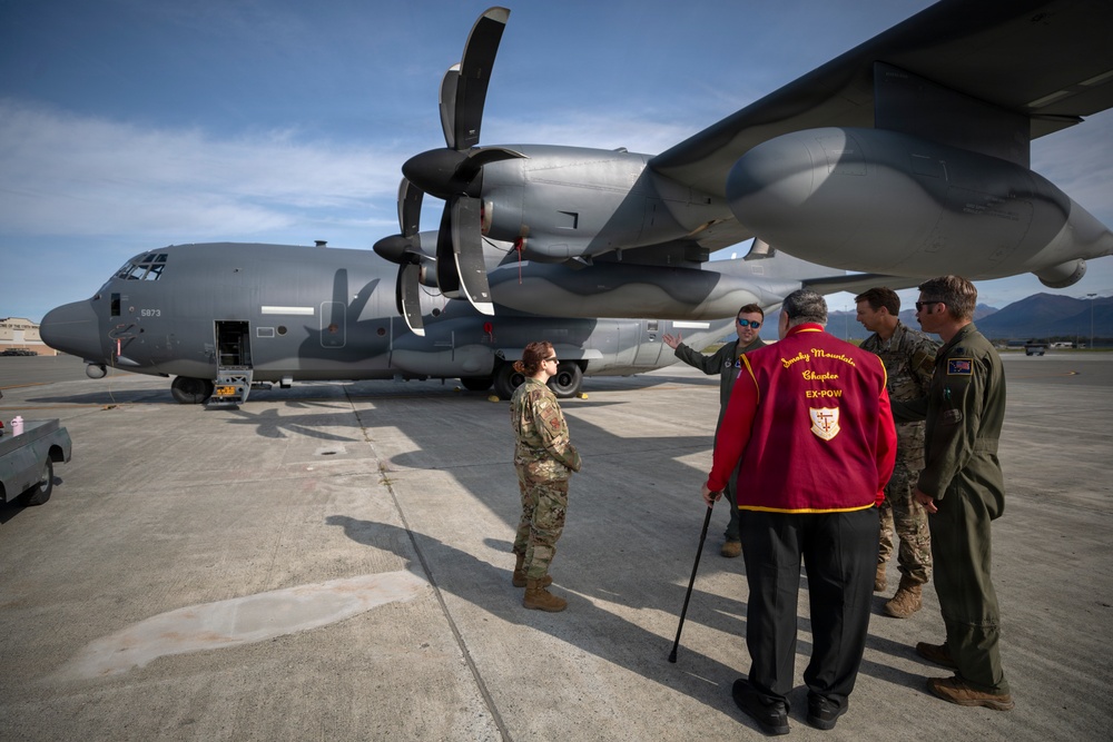 A group of military personnel and a civilian veteran are gathered on an airfield tarmac near a large military transport aircraft, likely a C-130 Hercules with tail number 5873. The civilian, wearing a red jacket identifying him as a member of the 'Smoky Mountains Chapter EX-POW' organization, appears to be receiving a briefing or tour of the aircraft. The setting appears to be a military air base with mountains visible in the background.
