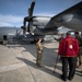 A group of military personnel and a civilian veteran are gathered on an airfield tarmac near a large military transport aircraft, likely a C-130 Hercules with tail number 5873. The civilian, wearing a red jacket identifying him as a member of the 'Smoky Mountains Chapter EX-POW' organization, appears to be receiving a briefing or tour of the aircraft. The setting appears to be a military air base with mountains visible in the background.
