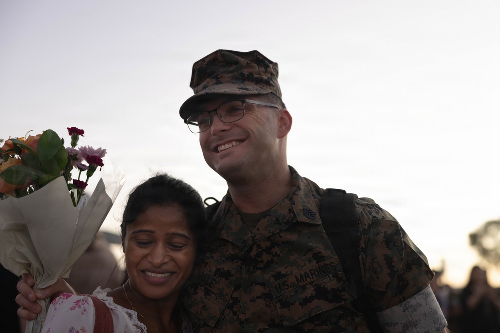 A U.S. Marine in MARPAT camouflage uniform is posing with a woman who is holding a bouquet of flowers. Both individuals are smiling, suggesting a joyful reunion, likely a homecoming event. The setting appears to be outdoors during golden hour lighting.