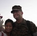 A U.S. Marine in MARPAT camouflage uniform is posing with a woman who is holding a bouquet of flowers. Both individuals are smiling, suggesting a joyful reunion, likely a homecoming event. The setting appears to be outdoors during golden hour lighting.