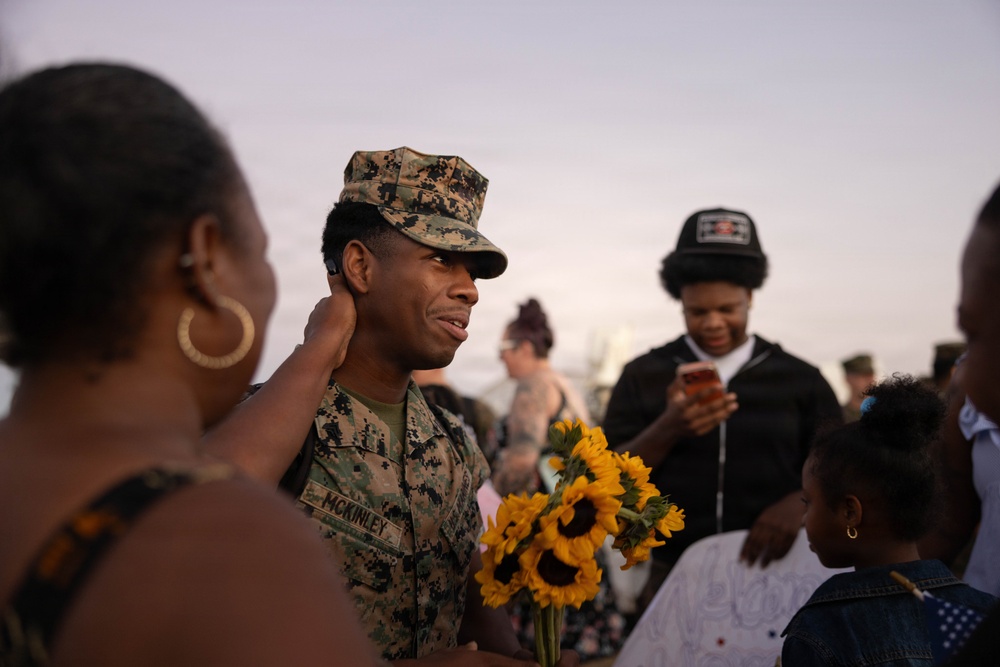A U.S. Marine in woodland MARPAT camouflage uniform, identified by the name tape 'McKINLEY,' is being greeted by family members during what appears to be a homecoming or graduation ceremony. He is holding a bouquet of sunflowers while a woman touches his face affectionately. The scene takes place outdoors at dusk with other people visible in the background.