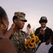 A U.S. Marine in woodland MARPAT camouflage uniform, identified by the name tape 'McKINLEY,' is being greeted by family members during what appears to be a homecoming or graduation ceremony. He is holding a bouquet of sunflowers while a woman touches his face affectionately. The scene takes place outdoors at dusk with other people visible in the background.