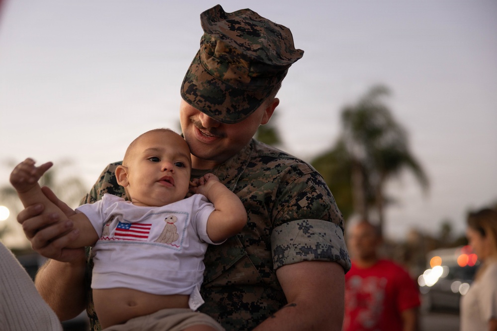 A U.S. Marine in MARPAT digital camouflage uniform holds a young infant dressed in a white onesie featuring an American flag embroidery. The Marine is smiling and leaning close to the baby in what appears to be an emotional reunion or family gathering at dusk. The setting appears to be outdoors, likely on or near a military base.