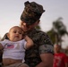 A U.S. Marine in MARPAT digital camouflage uniform holds a young infant dressed in a white onesie featuring an American flag embroidery. The Marine is smiling and leaning close to the baby in what appears to be an emotional reunion or family gathering at dusk. The setting appears to be outdoors, likely on or near a military base.