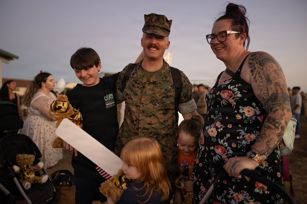 A U.S. Marine in woodland MARPAT camouflage uniform is reuniting with his family, likely at a homecoming event on a military base. He is surrounded by what appears to be his wife and three children, all smiling and embracing. The warm, golden-hour lighting and crowd in the background suggest a formal military homecoming ceremony.