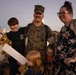 A U.S. Marine in woodland MARPAT camouflage uniform is reuniting with his family, likely at a homecoming event on a military base. He is surrounded by what appears to be his wife and three children, all smiling and embracing. The warm, golden-hour lighting and crowd in the background suggest a formal military homecoming ceremony.