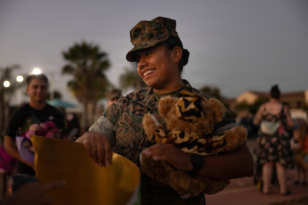 A female U.S. Marine in woodland MARPAT camouflage uniform is smiling broadly while holding a camouflage-dressed teddy bear and an envelope, likely during a homecoming or welcome ceremony. The setting appears to be an outdoor military base area at dusk, with other people visible in the blurred background. The scene conveys joy and emotional reunion.
