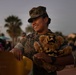 A female U.S. Marine in woodland MARPAT camouflage uniform is smiling broadly while holding a camouflage-dressed teddy bear and an envelope, likely during a homecoming or welcome ceremony. The setting appears to be an outdoor military base area at dusk, with other people visible in the blurred background. The scene conveys joy and emotional reunion.