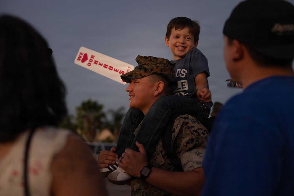A U.S. Marine in camouflage uniform and cover (cap) carries a young smiling child on his shoulders at what appears to be a military homecoming or welcome event. The child holds a white foam sign reading 'WE ❤ OUR HEROES.' The scene is set outdoors at dusk with other attendees visible in the foreground and background.