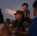 A U.S. Marine in camouflage uniform and cover (cap) carries a young smiling child on his shoulders at what appears to be a military homecoming or welcome event. The child holds a white foam sign reading 'WE ❤ OUR HEROES.' The scene is set outdoors at dusk with other attendees visible in the foreground and background.