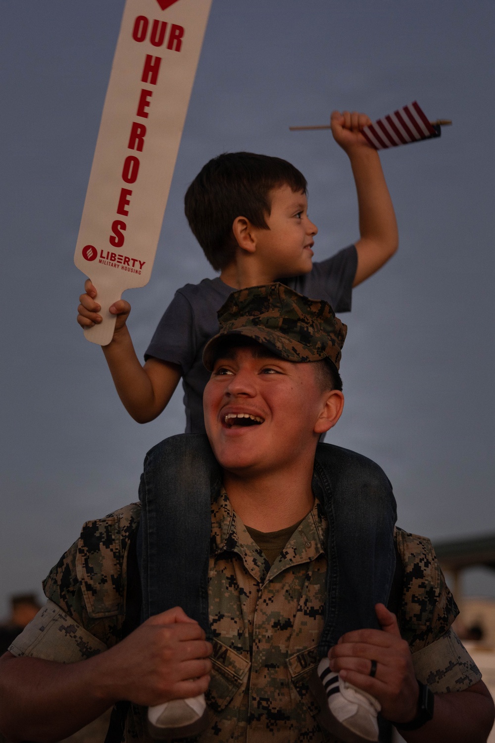 A smiling U.S. Marine in digital camouflage uniform and cover (cap) carries a young boy on his shoulders, likely during a homecoming or welcome-back event. The child holds a small American flag and a white sign reading 'OUR HEROES' sponsored by Liberty Military Housing. The scene conveys joy and celebration at what appears to be a military reunion event.