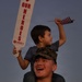 A smiling U.S. Marine in digital camouflage uniform and cover (cap) carries a young boy on his shoulders, likely during a homecoming or welcome-back event. The child holds a small American flag and a white sign reading 'OUR HEROES' sponsored by Liberty Military Housing. The scene conveys joy and celebration at what appears to be a military reunion event.