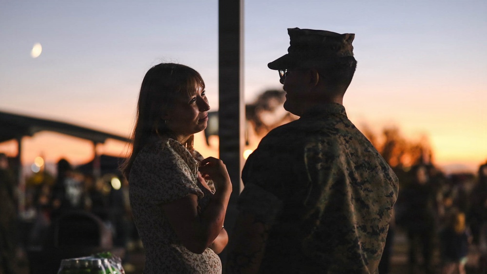 A woman in a floral dress and a person in military camouflage uniform face each other during twilight, appearing to be in conversation. The scene is backlit by a warm sunset sky with a visible moon, creating a partially silhouetted effect. Other people and structures are visible in the blurred background, suggesting a public or base gathering.