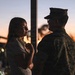 A woman in a floral dress and a person in military camouflage uniform face each other during twilight, appearing to be in conversation. The scene is backlit by a warm sunset sky with a visible moon, creating a partially silhouetted effect. Other people and structures are visible in the blurred background, suggesting a public or base gathering.