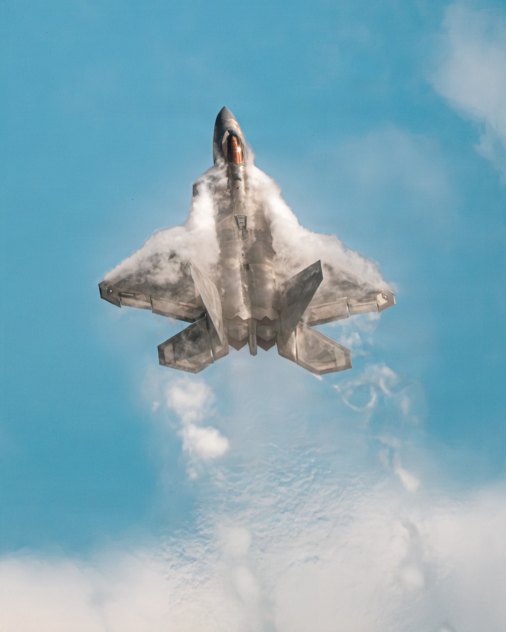 A fifth-generation stealth fighter jet, consistent with an F-22 Raptor, is photographed from below in a near-vertical climb against a blue sky. The aircraft is generating dramatic vapor condensation clouds across its fuselage and wings, likely due to rapid pressure changes during a high-performance maneuver. The image appears to have been taken at an airshow or during a demonstration flight.