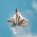 A fifth-generation stealth fighter jet, consistent with an F-22 Raptor, is photographed from below in a near-vertical climb against a blue sky. The aircraft is generating dramatic vapor condensation clouds across its fuselage and wings, likely due to rapid pressure changes during a high-performance maneuver. The image appears to have been taken at an airshow or during a demonstration flight.