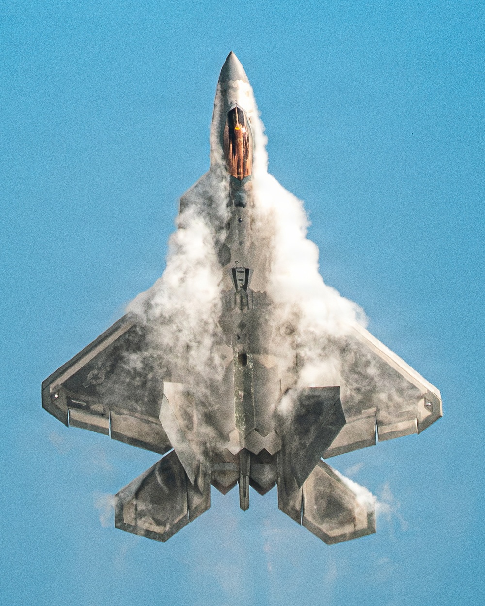 A fifth-generation stealth fighter jet, identifiable as an F-22 Raptor, is photographed from below in a near-vertical climb against a clear blue sky. The aircraft is generating a dramatic vapor cone or condensation cloud across its fuselage and wings, likely caused by rapid pressure changes during high-performance maneuvering. The afterburner glow is visible through the cockpit canopy area, indicating maximum thrust is being applied.