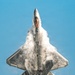 A fifth-generation stealth fighter jet, identifiable as an F-22 Raptor, is photographed from below in a near-vertical climb against a clear blue sky. The aircraft is generating a dramatic vapor cone or condensation cloud across its fuselage and wings, likely caused by rapid pressure changes during high-performance maneuvering. The afterburner glow is visible through the cockpit canopy area, indicating maximum thrust is being applied.