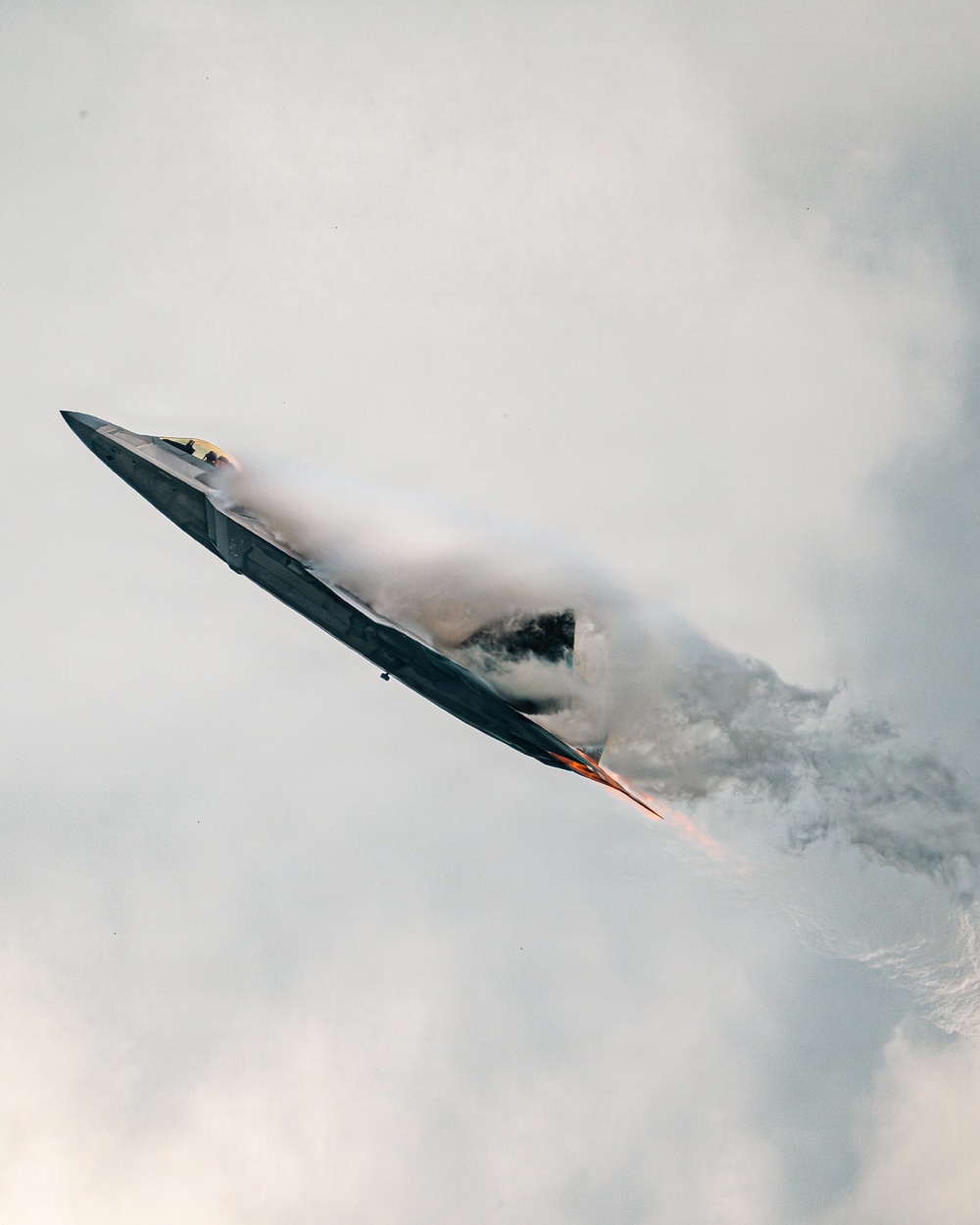 A stealth fighter jet, consistent with an F-22 Raptor, is captured in a steep climbing maneuver with afterburners fully engaged, producing visible orange flame at the exhaust nozzles. The aircraft is surrounded by a dense condensation cloud (vapor cone/vortex effect) generated by the high-speed, high-G maneuver in humid atmospheric conditions. This appears to be an airshow demonstration based on the dramatic performance profile.