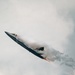 A stealth fighter jet, consistent with an F-22 Raptor, is captured in a steep climbing maneuver with afterburners fully engaged, producing visible orange flame at the exhaust nozzles. The aircraft is surrounded by a dense condensation cloud (vapor cone/vortex effect) generated by the high-speed, high-G maneuver in humid atmospheric conditions. This appears to be an airshow demonstration based on the dramatic performance profile.