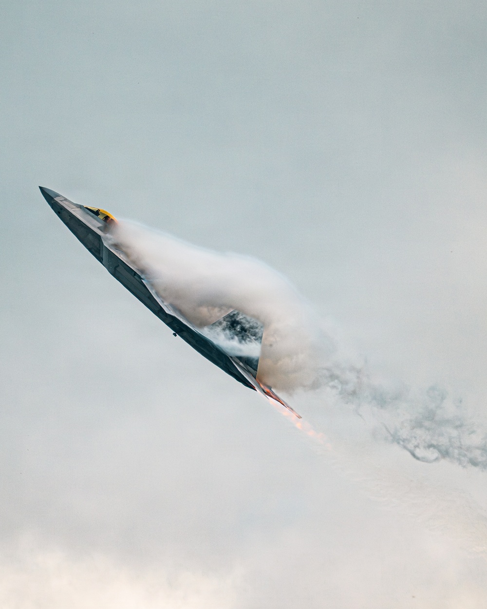 A stealth fighter jet, consistent with an F-22 Raptor, is captured in a steep climbing maneuver against an overcast sky. The aircraft is generating a dramatic vapor cone (condensation cloud) around its fuselage, indicative of high-speed, high-angle-of-attack flight. Afterburner flame is visible at the engine exhaust, and a dark smoke trail follows behind the aircraft.