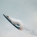 A stealth fighter jet, consistent with an F-22 Raptor, is captured in a steep climbing maneuver against an overcast sky. The aircraft is generating a dramatic vapor cone (condensation cloud) around its fuselage, indicative of high-speed, high-angle-of-attack flight. Afterburner flame is visible at the engine exhaust, and a dark smoke trail follows behind the aircraft.
