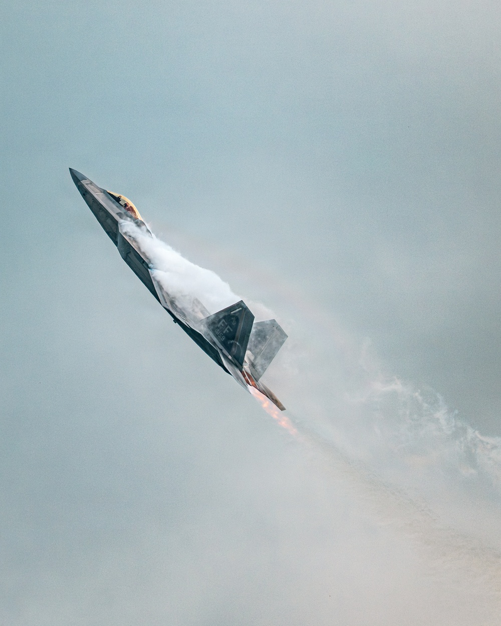 A U.S. Air Force F-22 Raptor stealth fighter jet is captured in a steep near-vertical climb during what appears to be an airshow demonstration. The aircraft is in full afterburner with visible engine flames and a dramatic vapor/condensation cloud forming around the fuselage due to rapid pressure changes. The tail code 'FF' and partial serial number are visible on the aircraft's body.