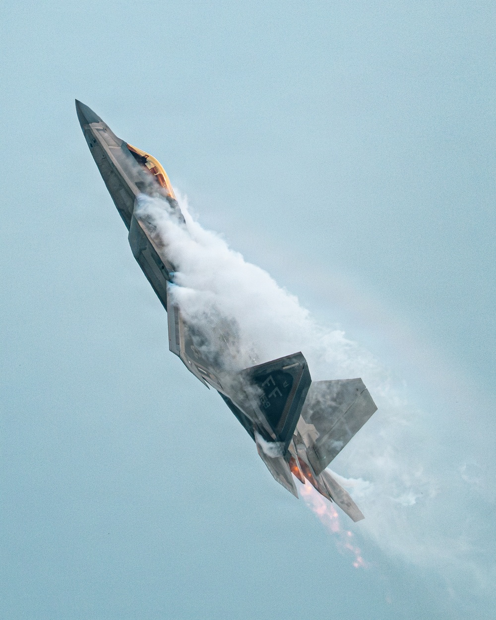 A Lockheed Martin F-22 Raptor fifth-generation stealth fighter jet is captured in a near-vertical climb maneuver against a pale blue-grey sky. The aircraft is producing a dramatic vapor/condensation cloud along its fuselage, consistent with a high-angle-of-attack maneuver at an airshow. Afterburner flame is visible at the engine exhausts, indicating full military or afterburner power.