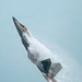 A Lockheed Martin F-22 Raptor fifth-generation stealth fighter jet is captured in a near-vertical climb maneuver against a pale blue-grey sky. The aircraft is producing a dramatic vapor/condensation cloud along its fuselage, consistent with a high-angle-of-attack maneuver at an airshow. Afterburner flame is visible at the engine exhausts, indicating full military or afterburner power.