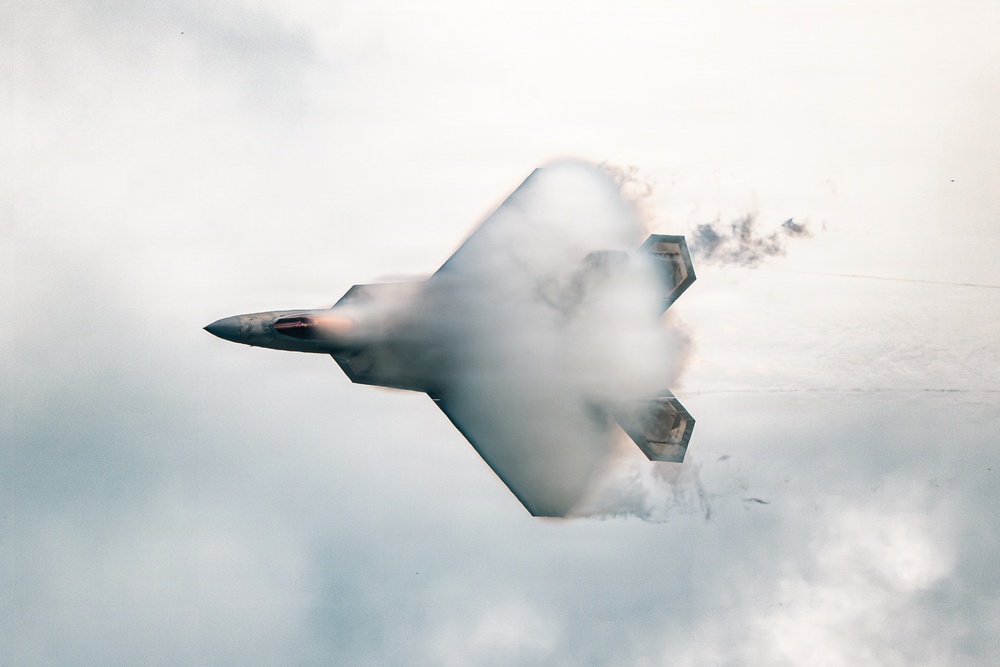 A stealth fighter jet, consistent with an F-22 Raptor design, is captured in a high-performance maneuver surrounded by a dense vapor cone and condensation cloud. The aircraft appears to be in a near-vertical or inverted banking attitude, with afterburner glow visible at the engine exhaust. The dramatic vapor effect suggests high-speed, high-G flight through humid air conditions.