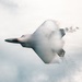 A stealth fighter jet, consistent with an F-22 Raptor design, is captured in a high-performance maneuver surrounded by a dense vapor cone and condensation cloud. The aircraft appears to be in a near-vertical or inverted banking attitude, with afterburner glow visible at the engine exhaust. The dramatic vapor effect suggests high-speed, high-G flight through humid air conditions.