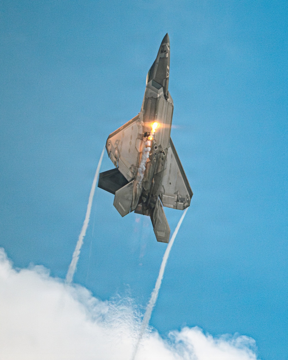 A fifth-generation stealth fighter jet, consistent with an F-22 Raptor, is captured in a near-vertical climb maneuver against a clear blue sky. The aircraft has its weapon bay doors open and afterburner ignited, producing a bright orange flame and smoke trail. Wingtip vortex condensation trails extend downward from the aircraft toward clouds below.
