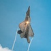 A fifth-generation stealth fighter jet, consistent with an F-22 Raptor, is captured in a near-vertical climb maneuver against a clear blue sky. The aircraft has its weapon bay doors open and afterburner ignited, producing a bright orange flame and smoke trail. Wingtip vortex condensation trails extend downward from the aircraft toward clouds below.