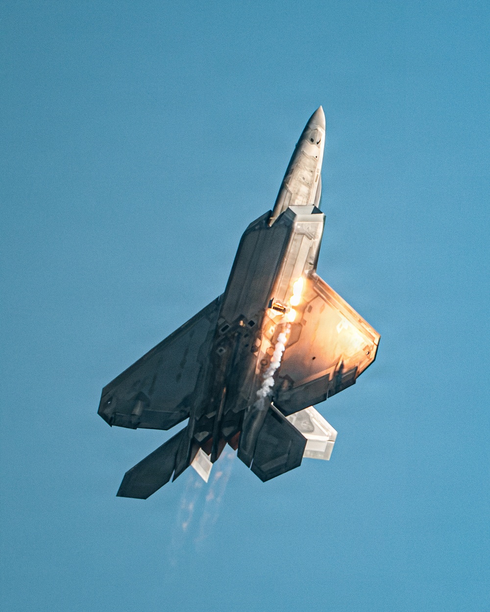 A fifth-generation stealth fighter jet, identifiable as an F-22 Raptor, is captured in a near-vertical climb maneuver against a clear blue sky. The aircraft is firing flares from its countermeasure dispensers, producing bright orange-white bursts of light and trailing smoke. This appears to be an airshow demonstration showcasing the aircraft's agility and defensive systems.