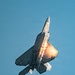 A fifth-generation stealth fighter jet, identifiable as an F-22 Raptor, is captured in a near-vertical climb maneuver against a clear blue sky. The aircraft is firing flares from its countermeasure dispensers, producing bright orange-white bursts of light and trailing smoke. This appears to be an airshow demonstration showcasing the aircraft's agility and defensive systems.