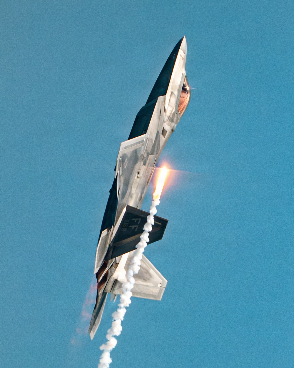 A military stealth fighter jet, identifiable as an F-22 Raptor, is captured in a near-vertical climb maneuver against a clear blue sky. The aircraft is dispensing flares and producing a visible afterburner flame plume, along with white condensation or smoke trails. Tail markings 'FF' are visible, indicating assignment to Langley Air Force Base's 1st Fighter Wing.