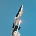 A military stealth fighter jet, identifiable as an F-22 Raptor, is captured in a near-vertical climb maneuver against a clear blue sky. The aircraft is dispensing flares and producing a visible afterburner flame plume, along with white condensation or smoke trails. Tail markings 'FF' are visible, indicating assignment to Langley Air Force Base's 1st Fighter Wing.
