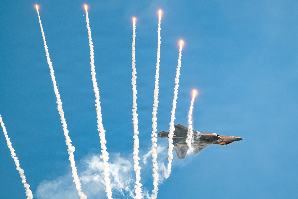 A military stealth fighter jet, consistent with an F-22 Raptor, is captured in a steep banking maneuver against a clear blue sky. The aircraft is simultaneously deploying multiple flares, which streak upward leaving bright white smoke trails. This appears to be an airshow demonstration showcasing the aircraft's countermeasure capabilities.