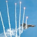A military stealth fighter jet, consistent with an F-22 Raptor, is captured in a steep banking maneuver against a clear blue sky. The aircraft is simultaneously deploying multiple flares, which streak upward leaving bright white smoke trails. This appears to be an airshow demonstration showcasing the aircraft's countermeasure capabilities.