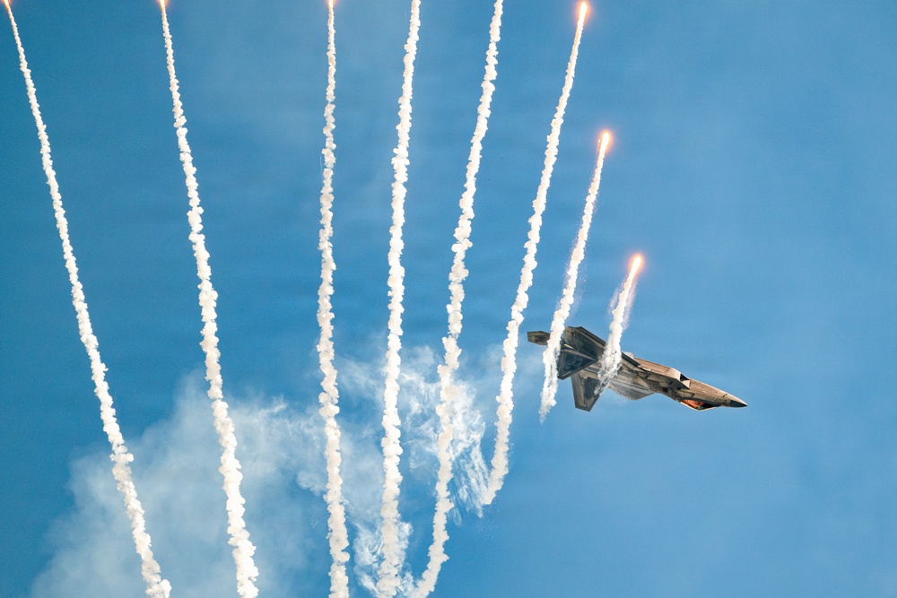 A military stealth fighter jet, consistent with an F-22 Raptor, is captured mid-maneuver against a clear blue sky while deploying multiple flares. The flares produce bright burning points and long white smoke trails that fan outward dramatically from the aircraft. This appears to be an airshow demonstration showcasing the aircraft's countermeasure systems.