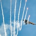 A military stealth fighter jet, consistent with an F-22 Raptor, is captured mid-maneuver against a clear blue sky while deploying multiple flares. The flares produce bright burning points and long white smoke trails that fan outward dramatically from the aircraft. This appears to be an airshow demonstration showcasing the aircraft's countermeasure systems.