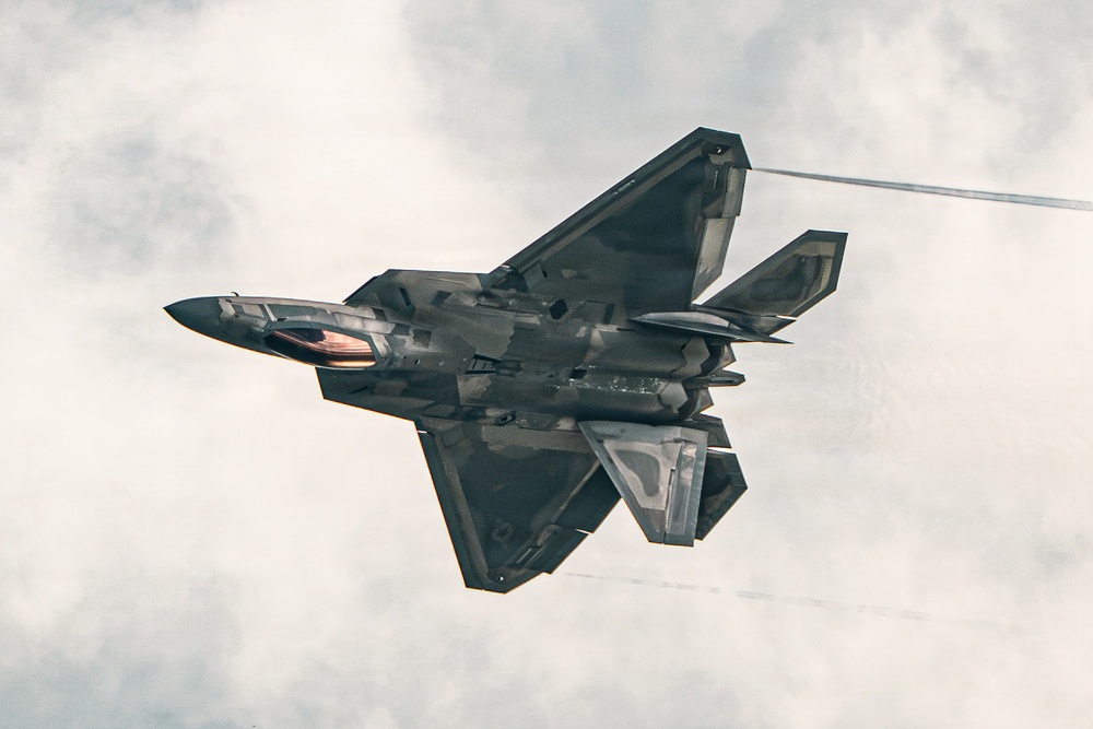 A dark-colored stealth fighter jet is captured in a banked maneuver from a below-and-behind perspective against an overcast sky. The aircraft displays characteristic angular stealth geometry with faceted surfaces, twin tail fins, and what appears to be an open weapons bay revealing internal components. Condensation trails or vapor streaks are visible trailing from the aircraft.