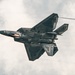 A dark-colored stealth fighter jet is captured in a banked maneuver from a below-and-behind perspective against an overcast sky. The aircraft displays characteristic angular stealth geometry with faceted surfaces, twin tail fins, and what appears to be an open weapons bay revealing internal components. Condensation trails or vapor streaks are visible trailing from the aircraft.