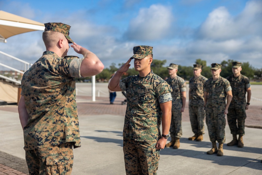 A group of U.S. Marines in MARPAT woodland camouflage uniforms are engaged in a formal saluting exchange on an outdoor parade deck or tarmac. Two Marines in the foreground are exchanging salutes face-to-face, while four additional Marines stand at attention in the background. The setting appears to be a military base, likely during a formal ceremony or inspection.