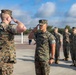 A group of U.S. Marines in MARPAT woodland camouflage uniforms are engaged in a formal saluting exchange on an outdoor parade deck or tarmac. Two Marines in the foreground are exchanging salutes face-to-face, while four additional Marines stand at attention in the background. The setting appears to be a military base, likely during a formal ceremony or inspection.