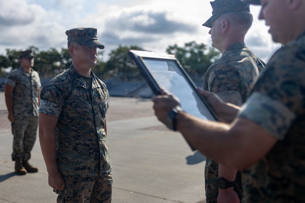 A military ceremony is taking place outdoors on a paved surface, likely a parade deck or tarmac. A Marine in MARPAT camouflage uniform stands at attention while another Marine presents a framed certificate or award document. Additional Marines are visible standing in the background.