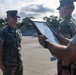 A military ceremony is taking place outdoors on a paved surface, likely a parade deck or tarmac. A Marine in MARPAT camouflage uniform stands at attention while another Marine presents a framed certificate or award document. Additional Marines are visible standing in the background.