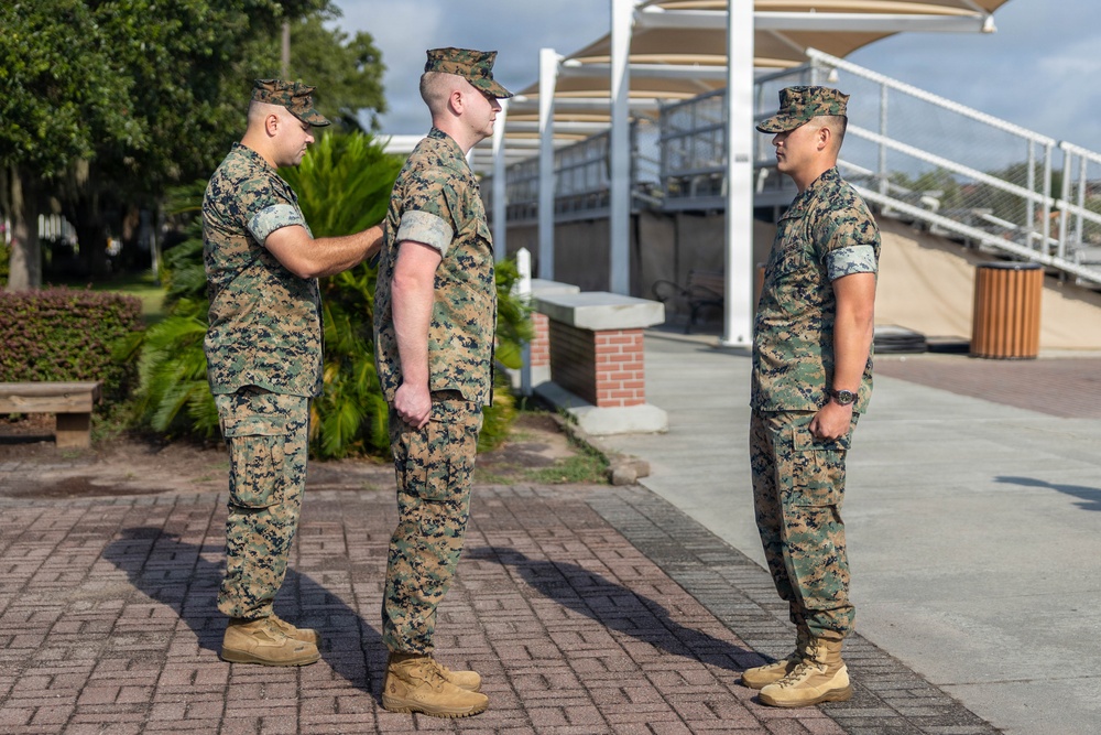 Three U.S. Marines in MARPAT woodland camouflage uniforms are gathered on a brick plaza in an outdoor military installation setting. One Marine on the left appears to be pinning an insignia or award onto the center Marine's uniform collar or chest area, suggesting a promotion or award ceremony. A third Marine stands at attention facing them, likely serving as a witness to the formal proceeding.